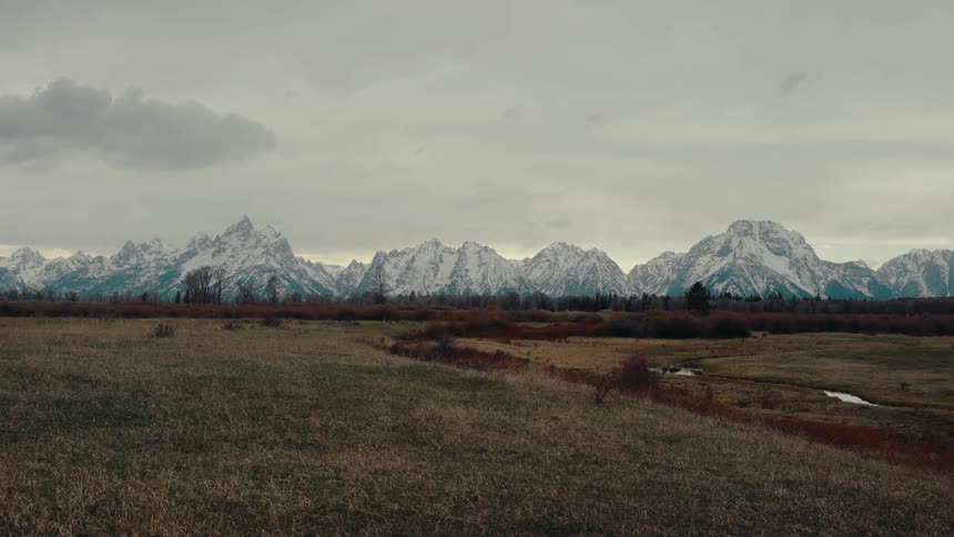 A vast grassy plain sits before the majestic, snow-capped Grand Teton mountain range on a cloudy, overcast day