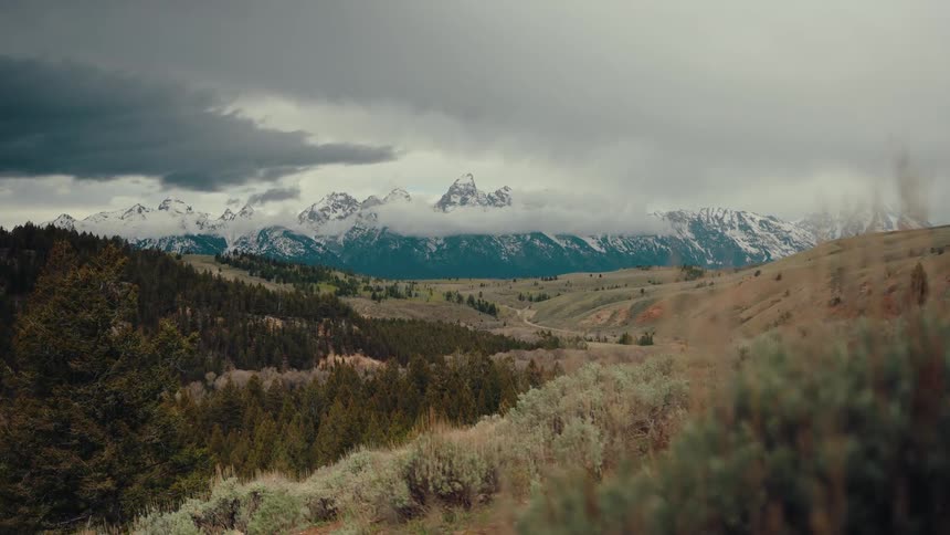 A wide shot of the majestic snow-capped Teton mountain range under a blanket of dark, stormy clouds, with rolling hills and evergreen forests in the foreground
