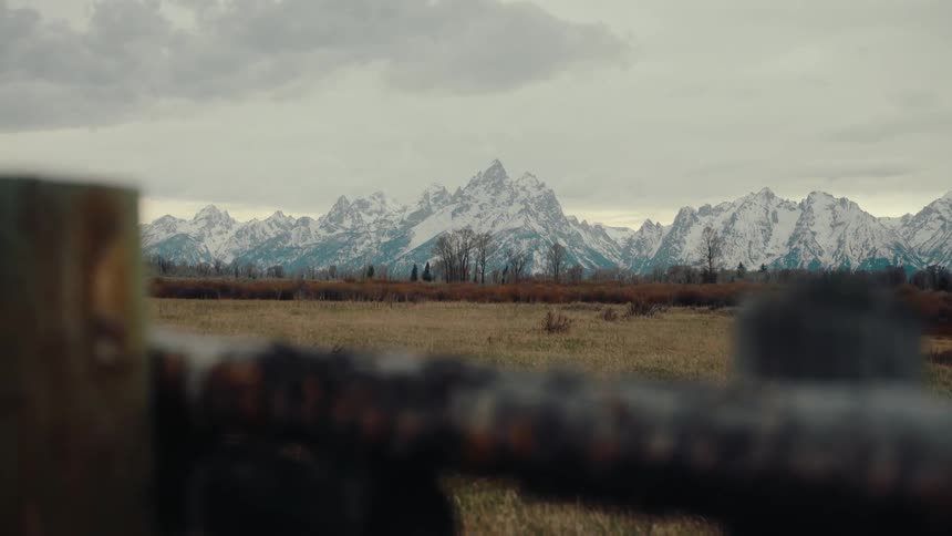 A static shot reveals the majestic snow-capped Teton Range from behind a rustic wooden fence on a cloudy day