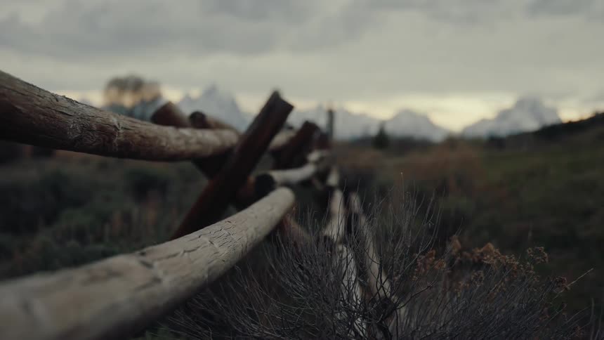 A rustic wooden fence stretches across a vast field towards a majestic, out-of-focus mountain range under a cloudy sky