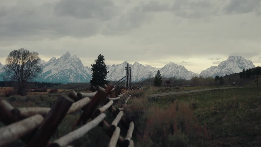 The majestic, snowy Grand Teton mountains stand tall behind a classic wooden ranch fence on an overcast day