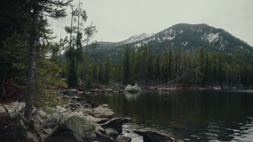 A tranquil mountain lake reflects the surrounding evergreen forest and snow-dusted peaks on an overcast day