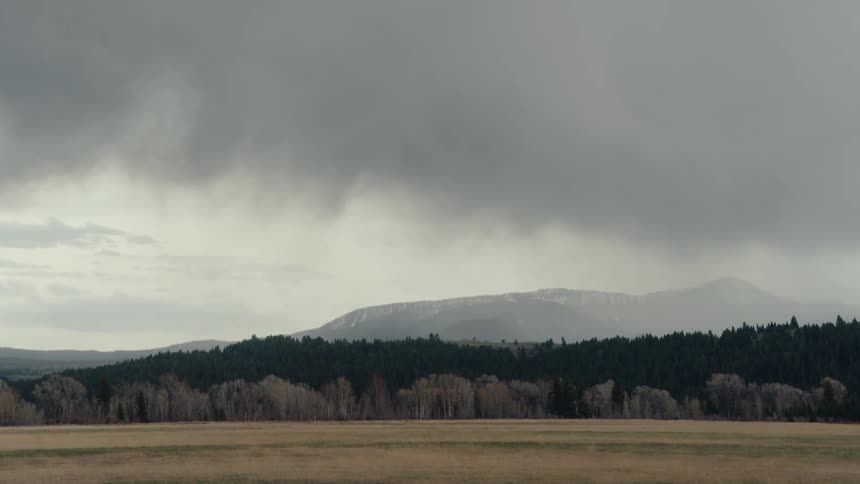 A moody timelapse captures a dramatic storm system passing over a wide-open field and forested, snow-capped mountains