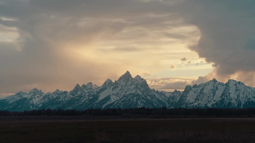 Dramatic storm clouds drift over the majestic snow-capped peaks of the Grand Teton mountain range during a golden sunset