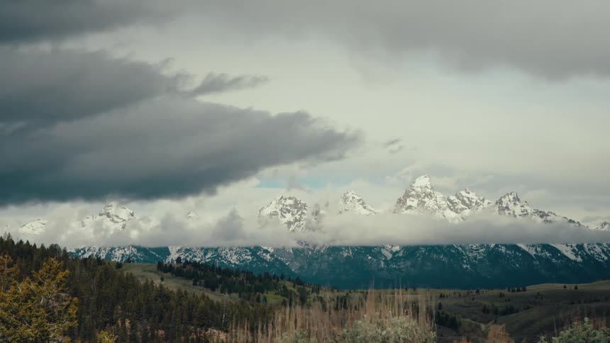 Dark storm clouds move in a dramatic timelapse across the sky, partially obscuring the magnificent snow-capped peaks of the Grand Teton mountain range