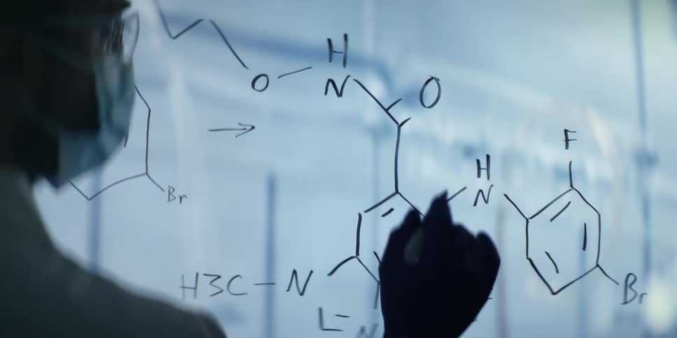 A scientist in a lab coat meticulously writes a complex chemical formula on a transparent board