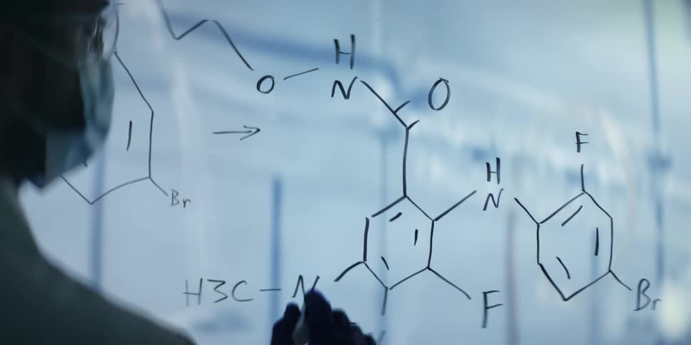 A scientist wearing gloves and a lab coat meticulously writes a complex chemical formula on a transparent board in a modern laboratory