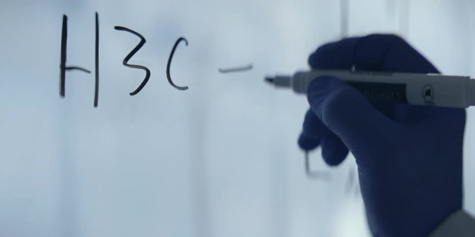 A chemist in a sterile lab carefully writes a complex chemical equation on a glass board with a marker