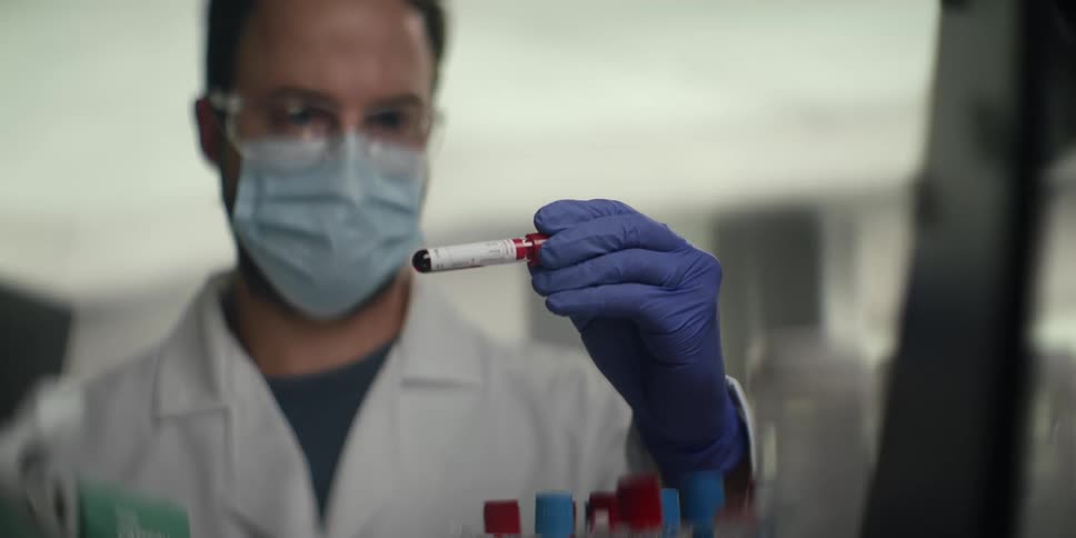 A focused male scientist in a lab coat and protective gear carefully examines a blood sample in a test tube