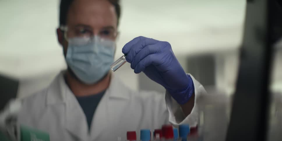 A male scientist wearing a mask and safety glasses carefully examines a clear liquid sample in a glass vial in a laboratory