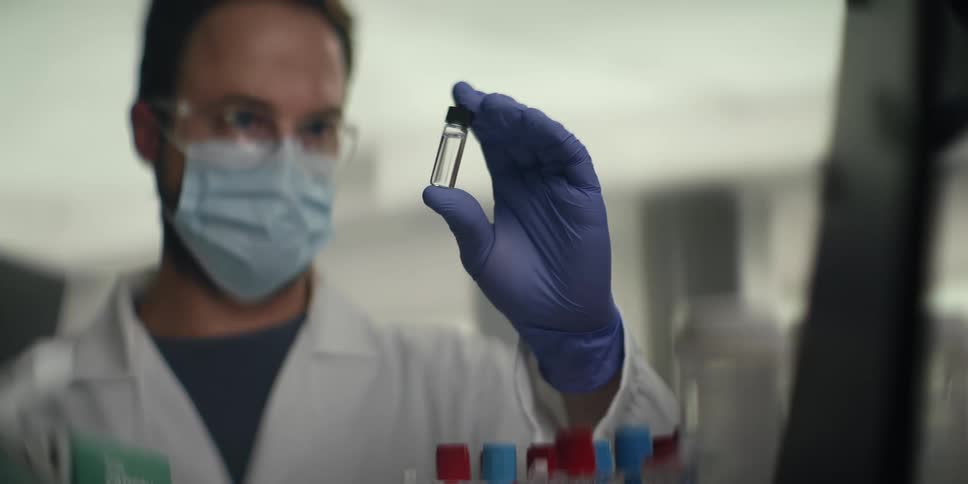 A focused scientist in a lab coat, mask, and gloves carefully examines liquid samples in test tubes