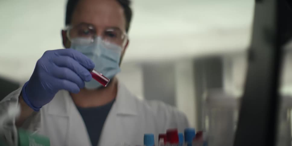 A male scientist wearing a lab coat, mask, and goggles carefully examines a vial of red liquid in a laboratory