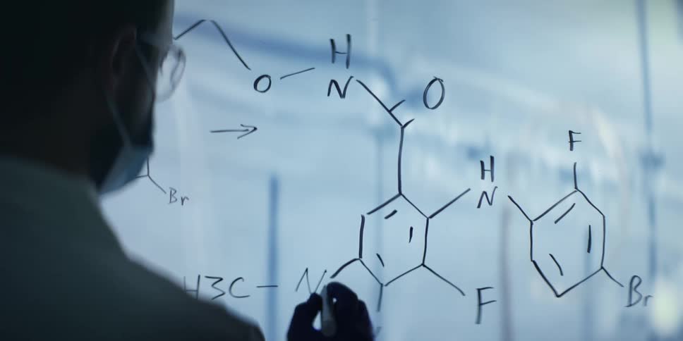 A scientist wearing a mask and gloves carefully writes a complex chemical formula on a glass board in a research lab