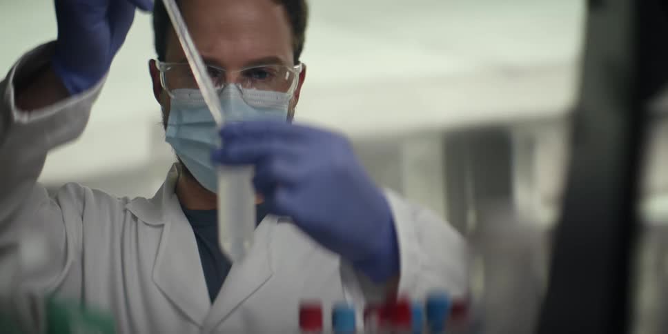 A focused scientist wearing a lab coat, mask, and safety glasses carefully transfers liquid with a pipette in a laboratory