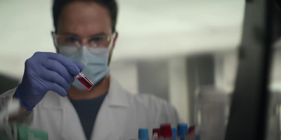 A focused male scientist wearing a lab coat and protective gear carefully examines a small vial of red liquid in a modern laboratory