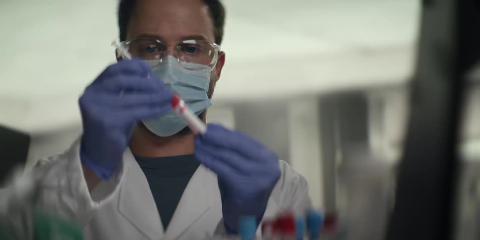 A male scientist wearing a lab coat, face mask, and gloves carefully handles a test tube with a red liquid sample in a laboratory