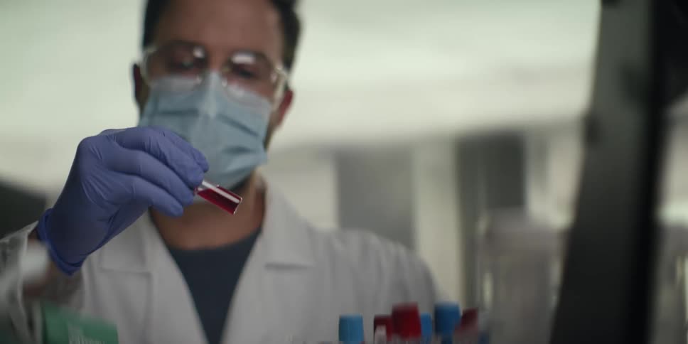 Focused male scientist in full protective gear meticulously examines a vial containing a red liquid sample in a research laboratory