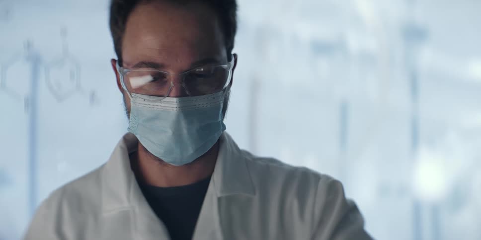 A male scientist wearing a mask and safety goggles in a laboratory closely examines a vial as the red liquid inside undergoes a chemical reaction and turns clear