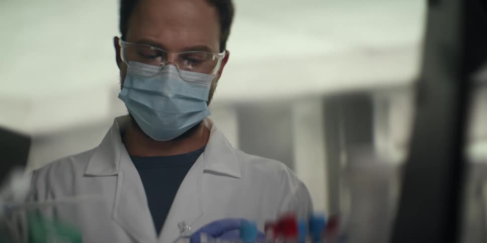 A focused male scientist in full protective gear carefully handles test tubes and uses a pipette in a modern laboratory