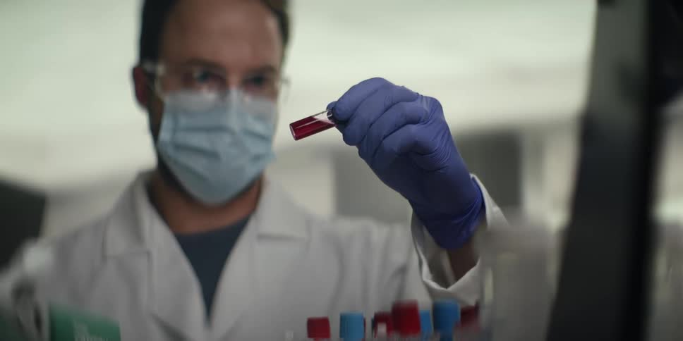 A male scientist wearing a lab coat, mask, and gloves carefully examines a test tube with a red liquid