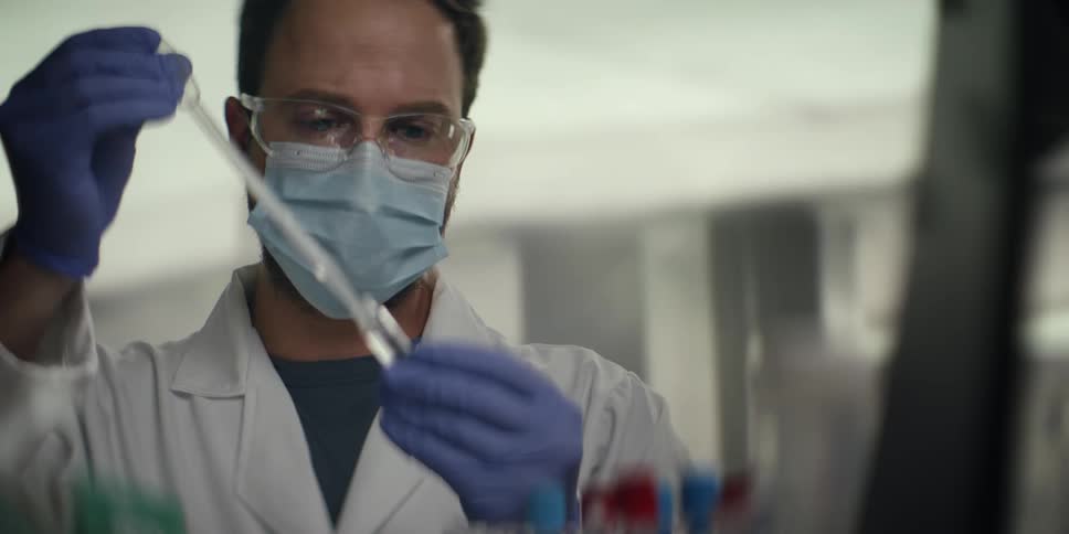 A male scientist in a lab coat and protective gear carefully uses a pipette to transfer liquid into a test tube