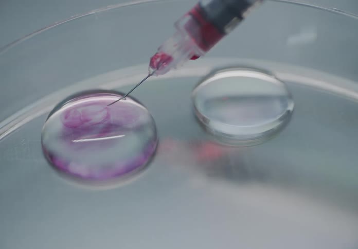 A syringe with a needle slowly injects a colored chemical into a droplet of clear liquid in a petri dish for a lab experiment