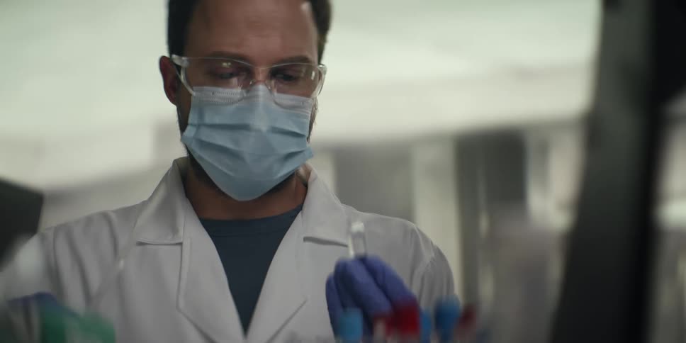 A male scientist wearing a mask and safety goggles carefully uses a pipette to transfer liquid into a test tube in a laboratory