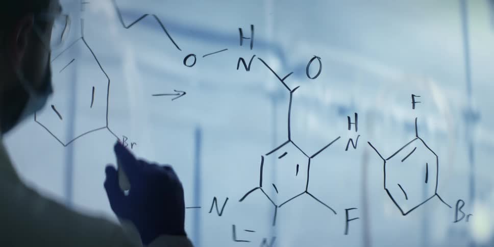 A chemist in protective gear meticulously works through a complex molecular formula on a transparent board in a modern laboratory