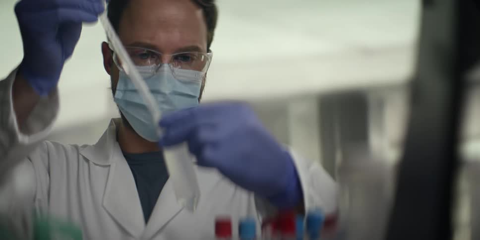 A male scientist in a lab coat and protective gear carefully handles a flask and pipette while working in a research laboratory