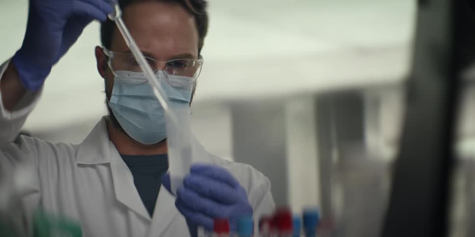 A focused male scientist in a lab coat and mask uses a pipette to conduct a medical experiment in a research laboratory