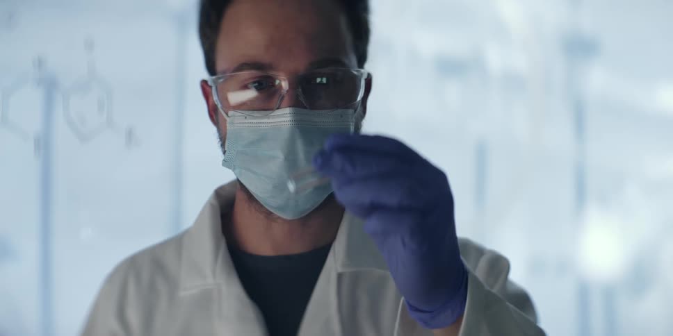 A male scientist in a protective mask and goggles carefully examines a red liquid sample in a vial before switching to a clear one in a modern laboratory