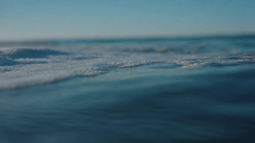 A serene, slow-motion, close-up view of the calm ocean surface with delicate sea foam under a clear blue sky