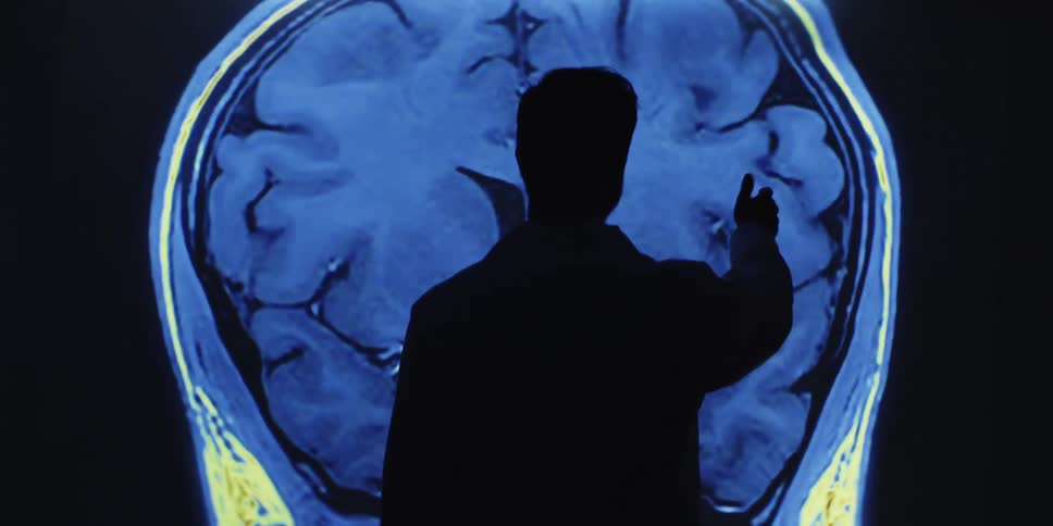A silhouetted doctor or scientist analyzes a colorful, animated MRI brain scan projected on a screen