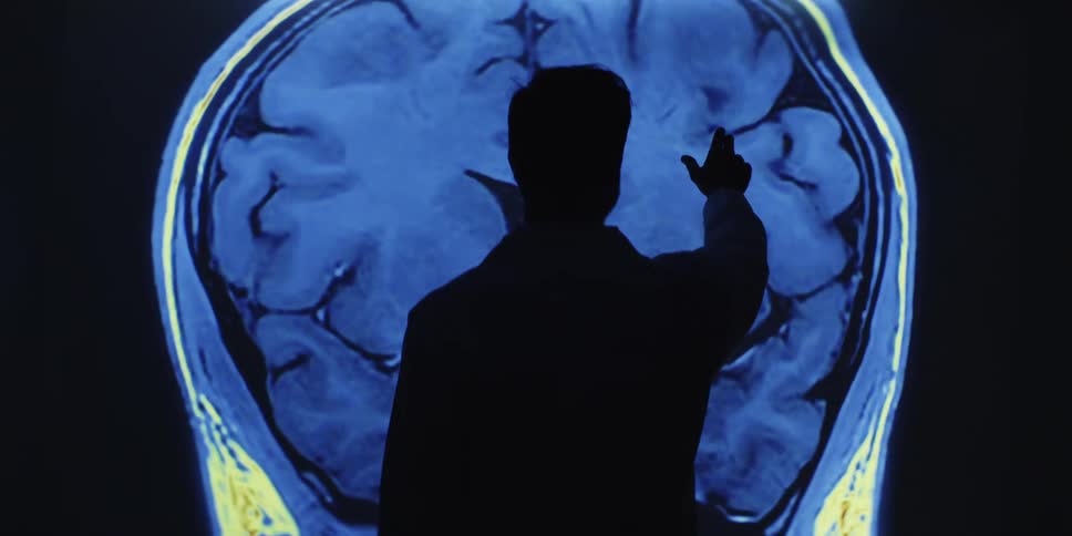 A silhouetted doctor gestures and points at a projected, animated MRI scan of a human brain