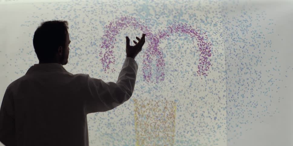 A male scientist in a lab coat manipulates a dynamic, colorful particle simulation on a large futuristic touch screen