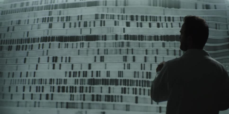 A male scientist in silhouette studies a massive wall projection of DNA sequencing results, gesturing as he analyzes the data