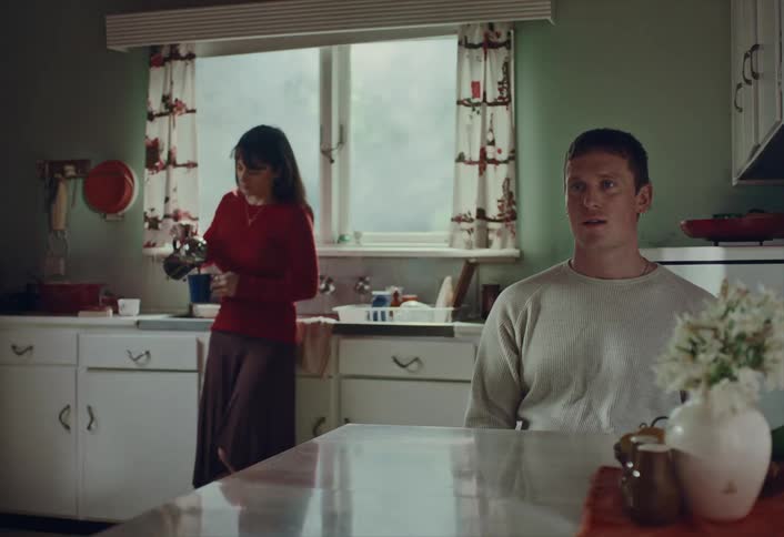 A man watches a woman prepare coffee in their sunlit vintage-style kitchen in the morning