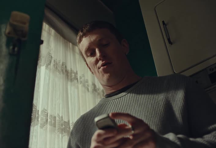 A young man nostalgically types a message on an old mobile phone, then looks out the window with a hopeful smile