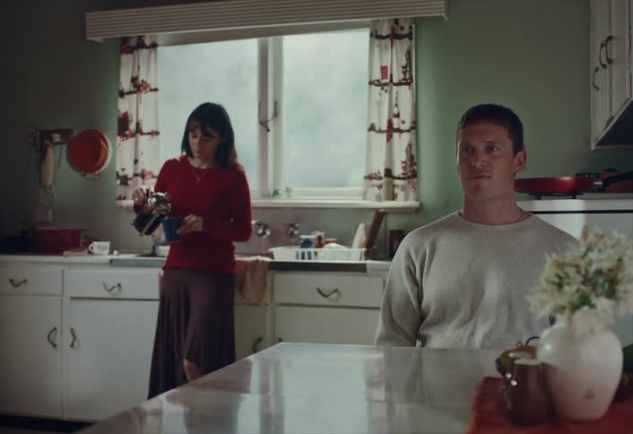 A contemplative man sits at the kitchen table in a vintage home while a woman prepares coffee in the background