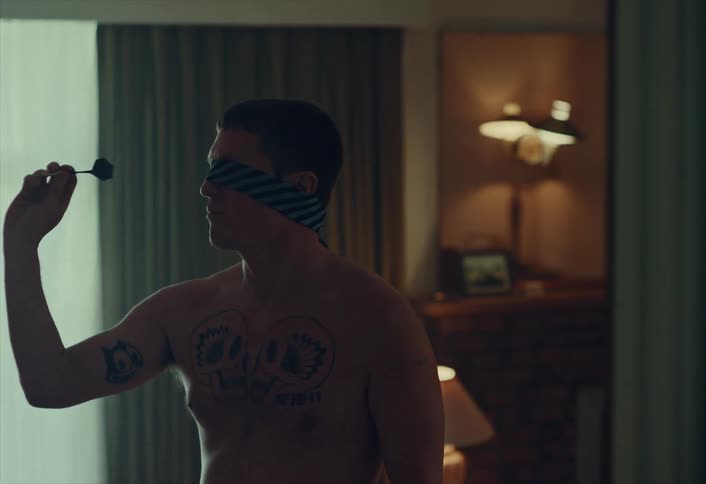 A shirtless tattooed man wearing a blindfold throws a dart in slow motion in a dimly lit room