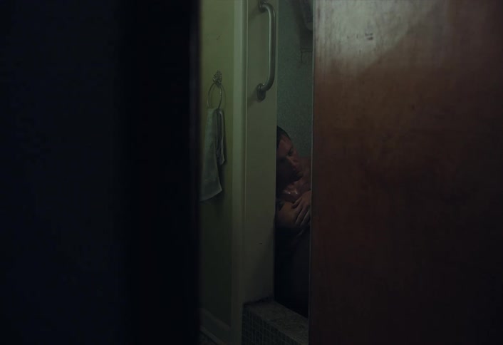 A lonely and depressed man sits in a dark, moody shower stall, viewed from behind a door