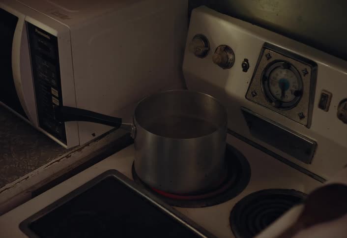 Water begins to boil in a metal pot on an old electric stove in a dark kitchen corner