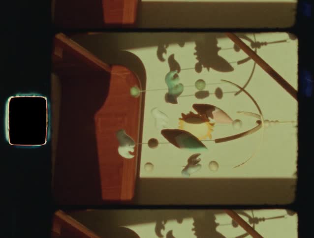 A vertical Super 8 film scan captures a whimsical, colorful hanging mobile as it gently sways and casts shadows on the wall