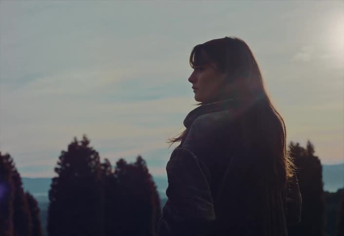A thoughtful woman stands silhouetted against the setting sun, gazing out at a serene landscape of trees and rolling hills