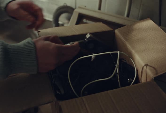 A person's hands sift through a cardboard box of old electronics and memories, picking up a vintage MP3 player