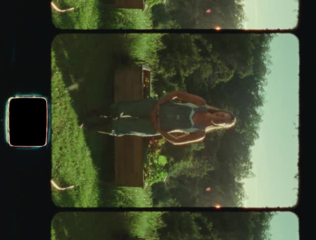 Vertical vintage film footage of a young woman in overalls standing and smiling in a lush green garden on a sunny day