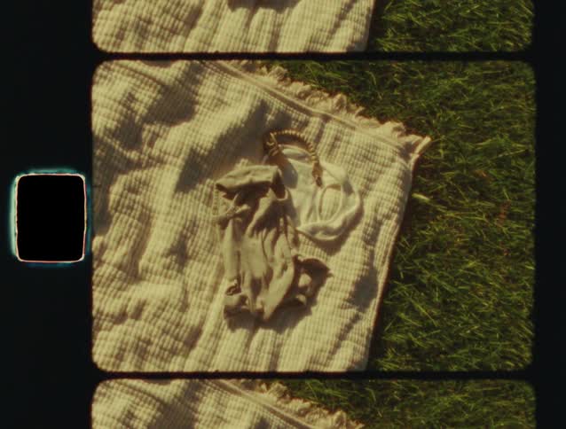 A baby's onesie and pacifier clip rest on a soft blanket in the sunlit grass in a nostalgic film clip