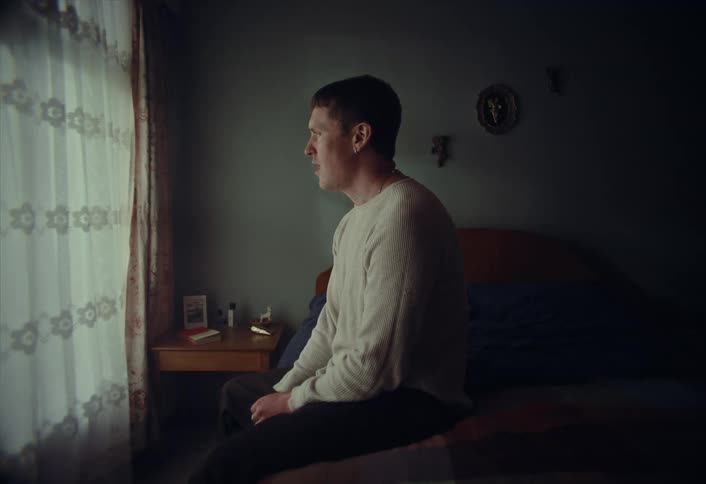 A young man sits alone in his dimly lit bedroom, looking pensively out the window while talking to himself