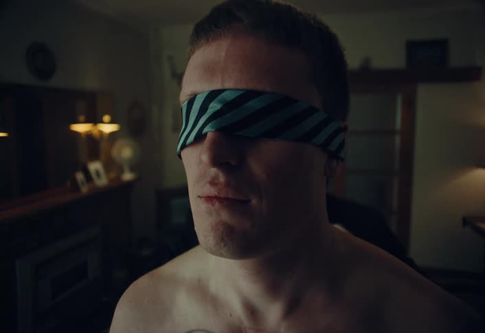 A shirtless young man is blindfolded with a striped necktie by another man in white gloves in a dimly lit, mysterious room
