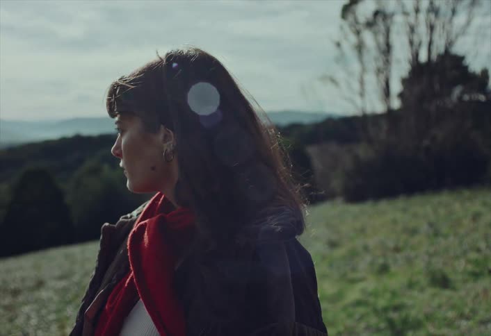 A thoughtful young woman with a red scarf stands in a windy, sunlit meadow before turning and walking out of frame